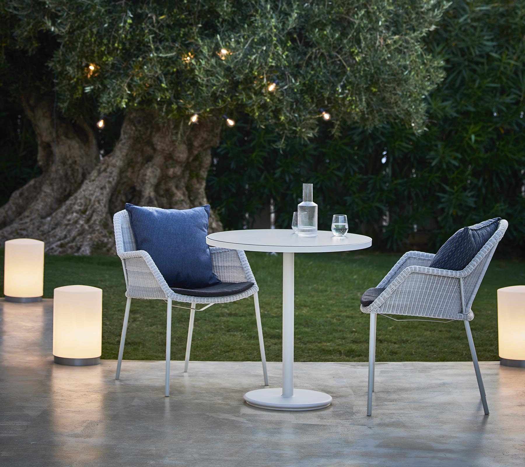 A cozy seating area featuring two modern chairs, a round table, and stylish lamps, surrounded by greenery and warm lighting.