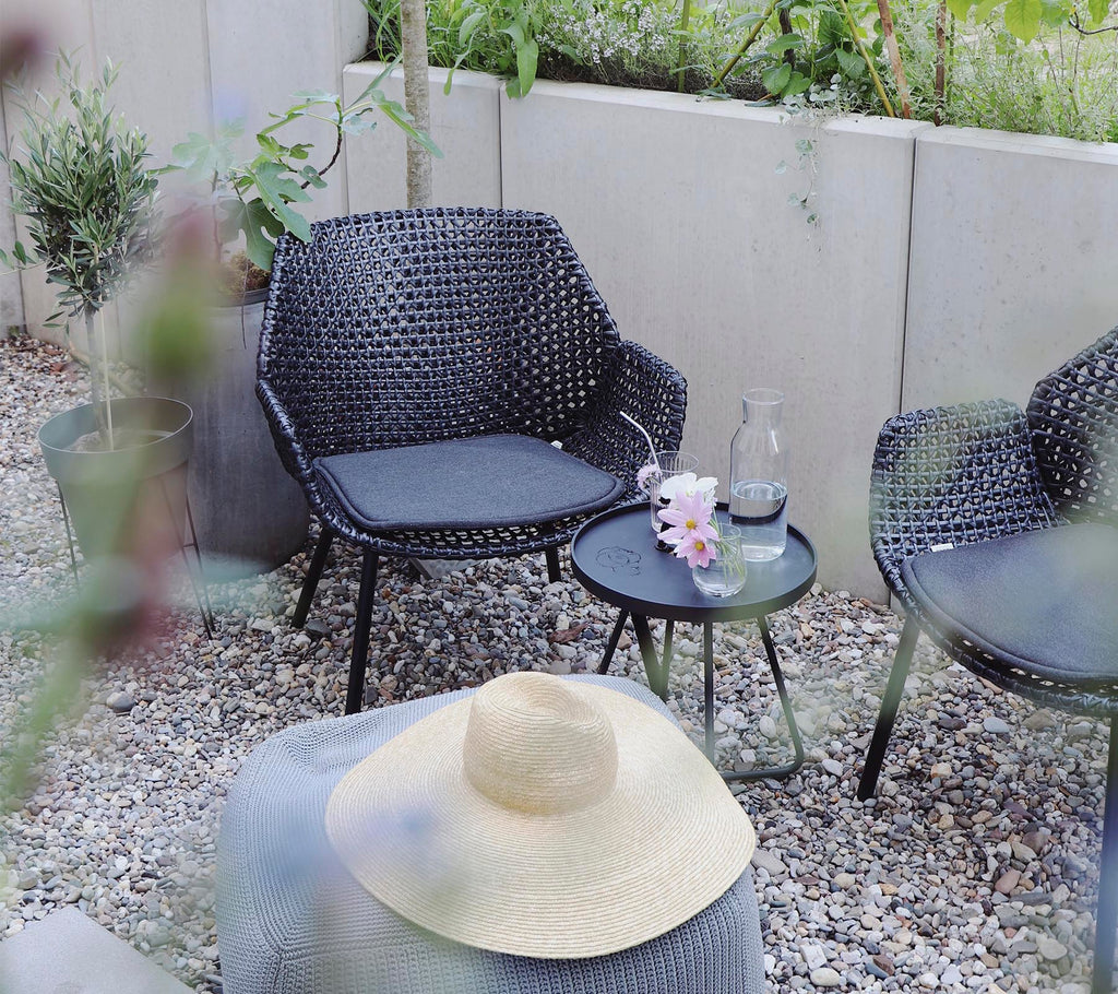A cozy seating area with woven chairs, a small table, a glass of water, flowers, and a straw hat, surrounded by greenery and stones.