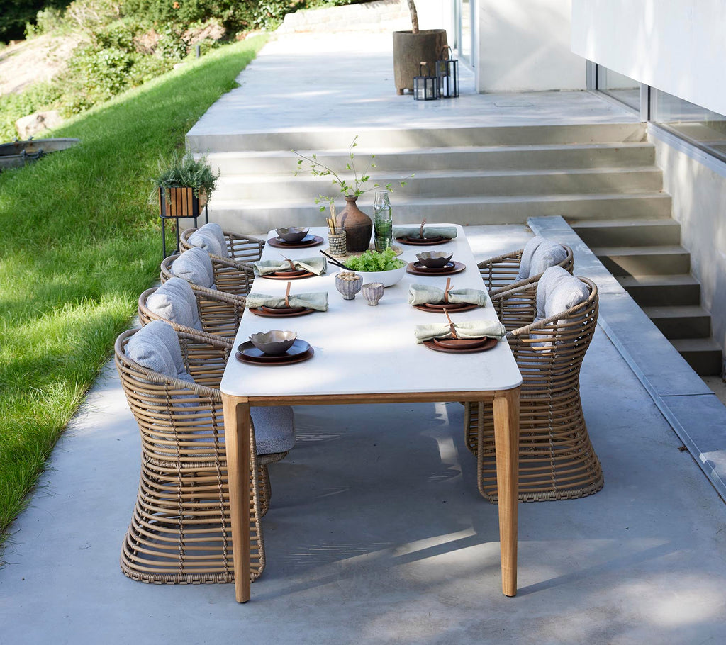 A stylish outdoor dining table set with round wicker chairs, elegantly arranged with plates and a centerpiece of greenery.
