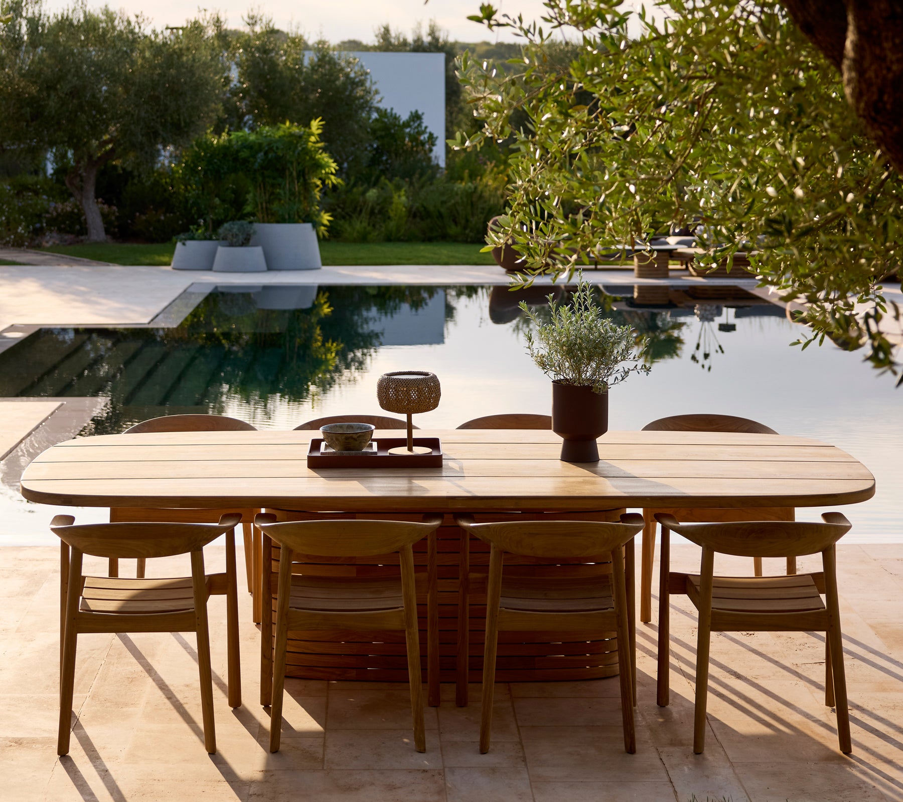 Modern wooden dining table and chairs set near a pool surrounded by greenery.