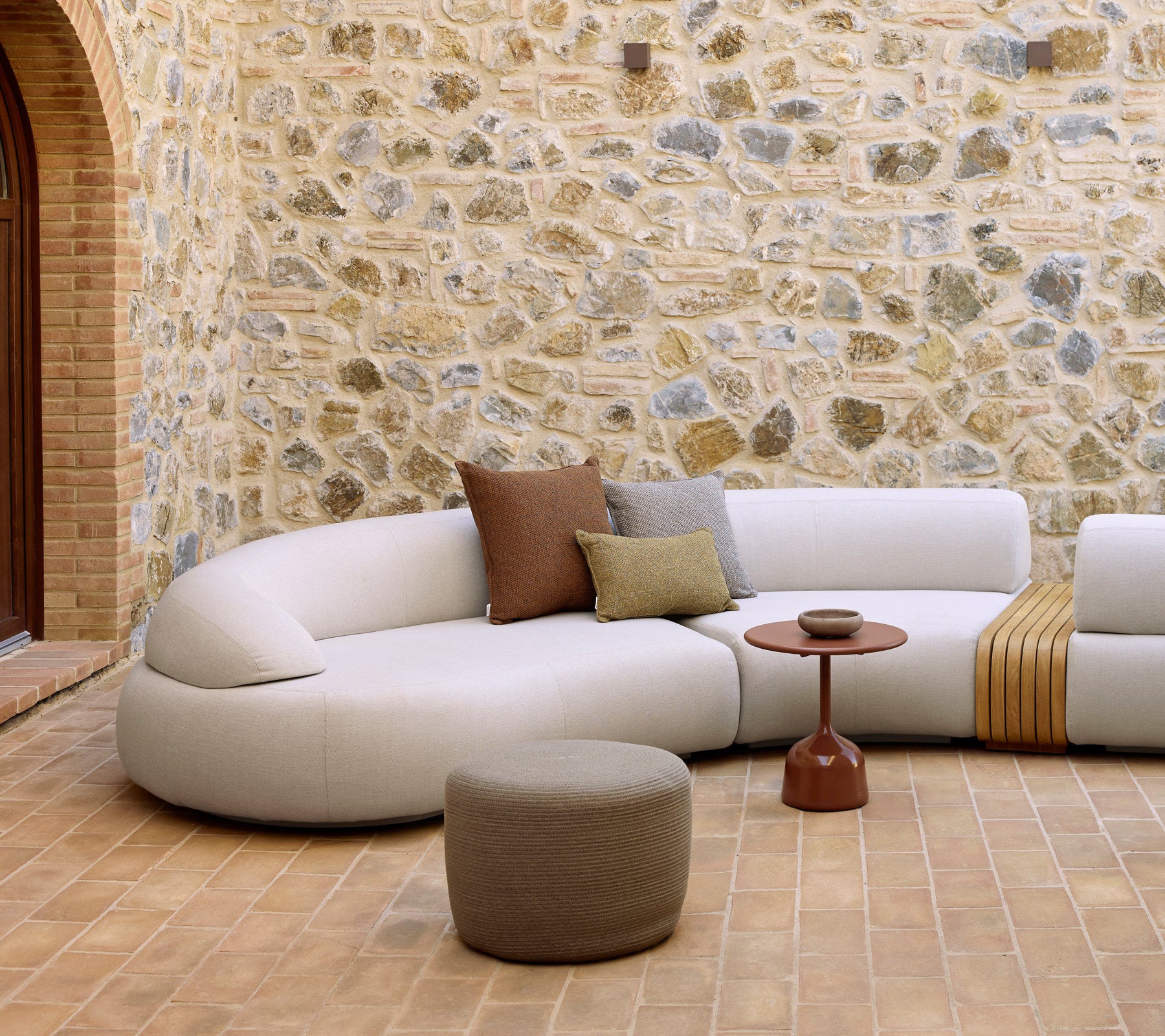Mellow lounge area featuring a curved sofa, Glaze coffee table, and a round footstool against a textured stone wall.