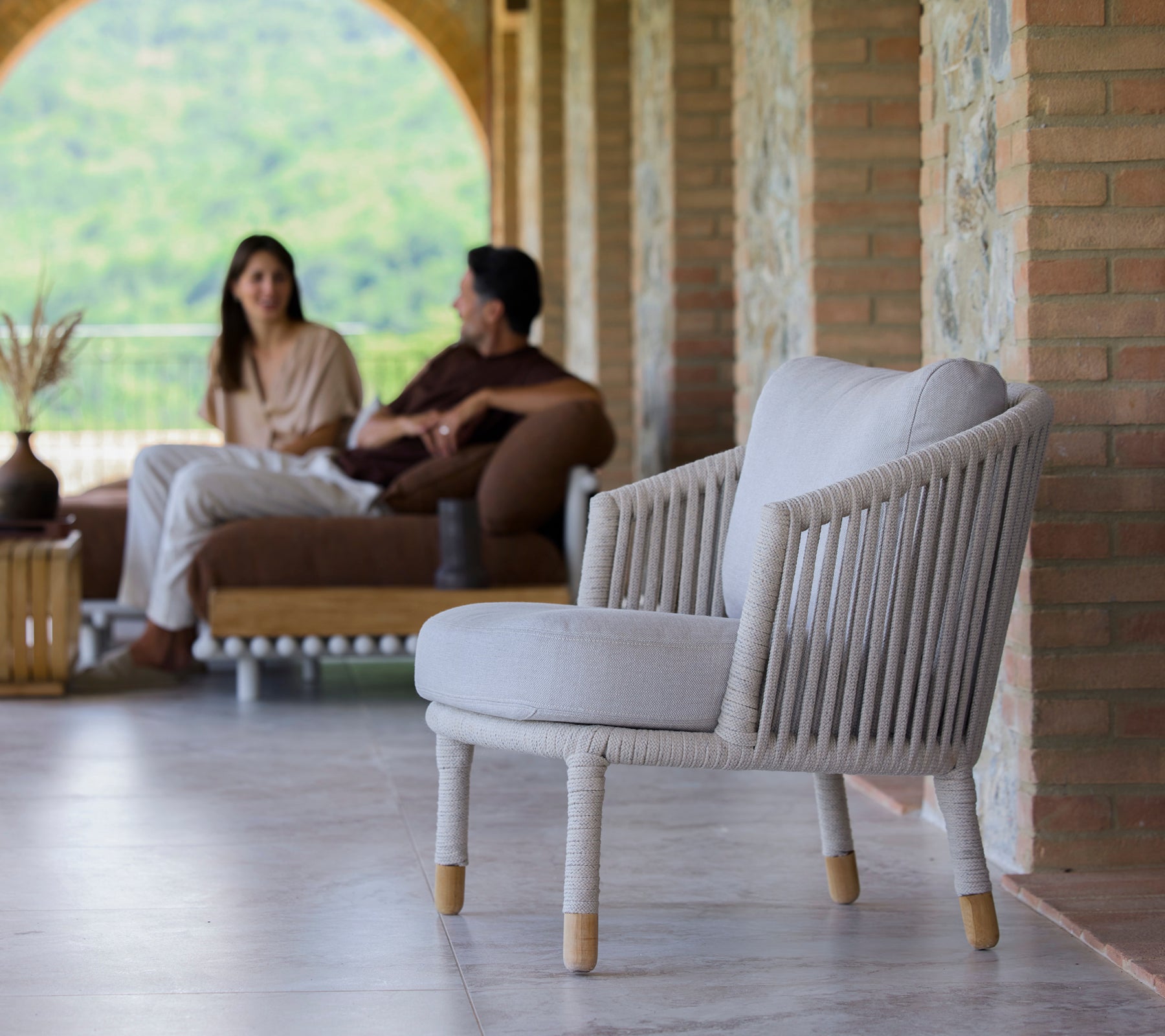 A sand-colored lounge chair with a minimalist design, featured in a serene setting with people engaged in conversation.