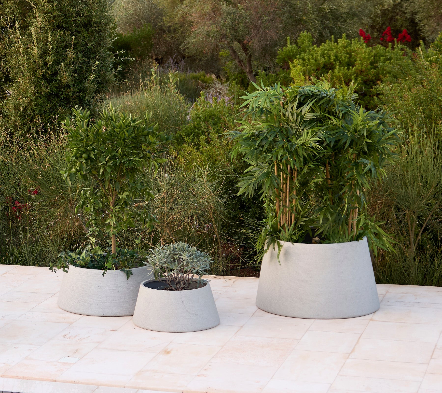 Three modern grey planters with various plants in a garden setting.
