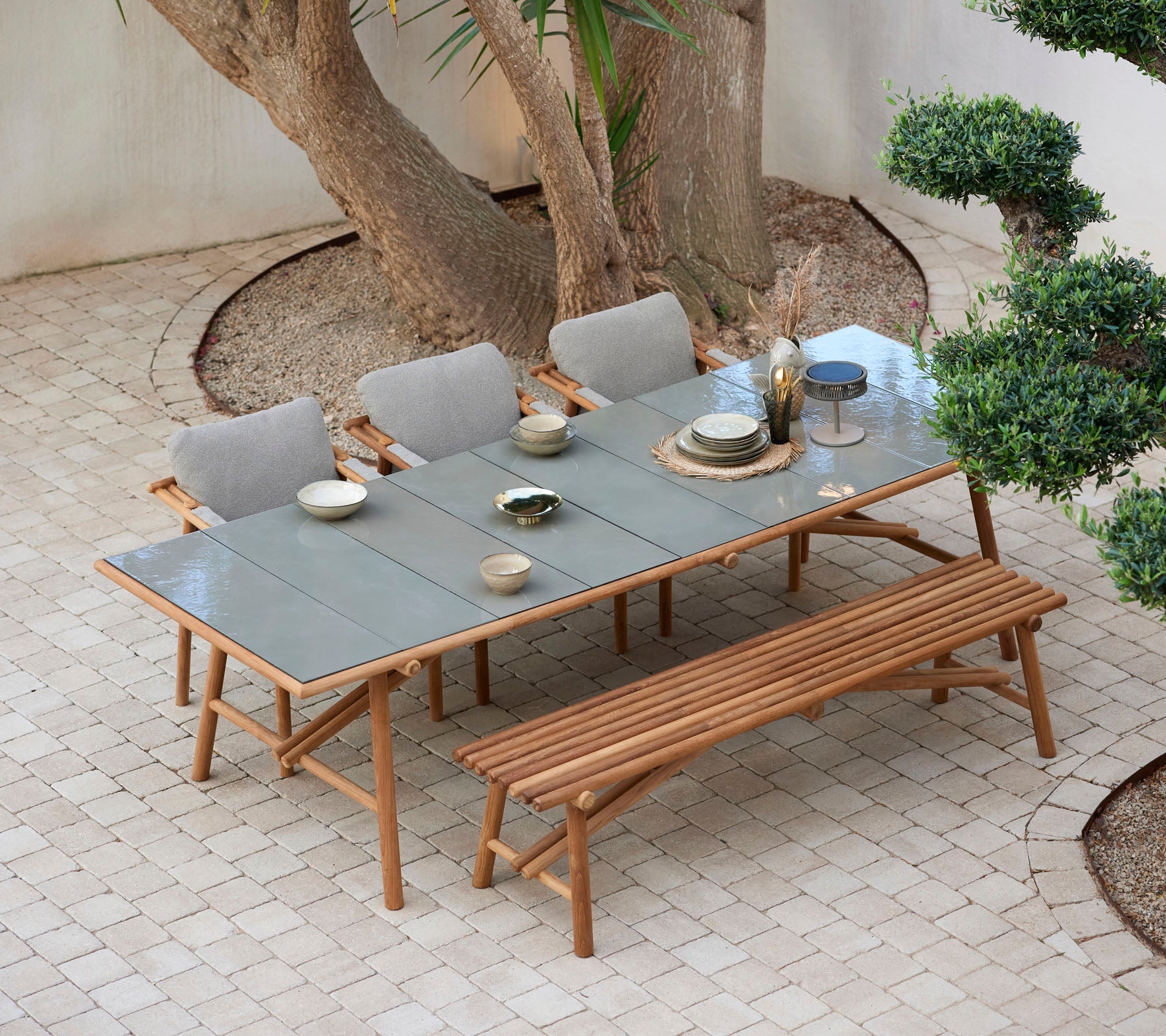 A wooden table set with dishes and glasses, surrounded by comfortable chairs and a bench, all positioned among greenery.