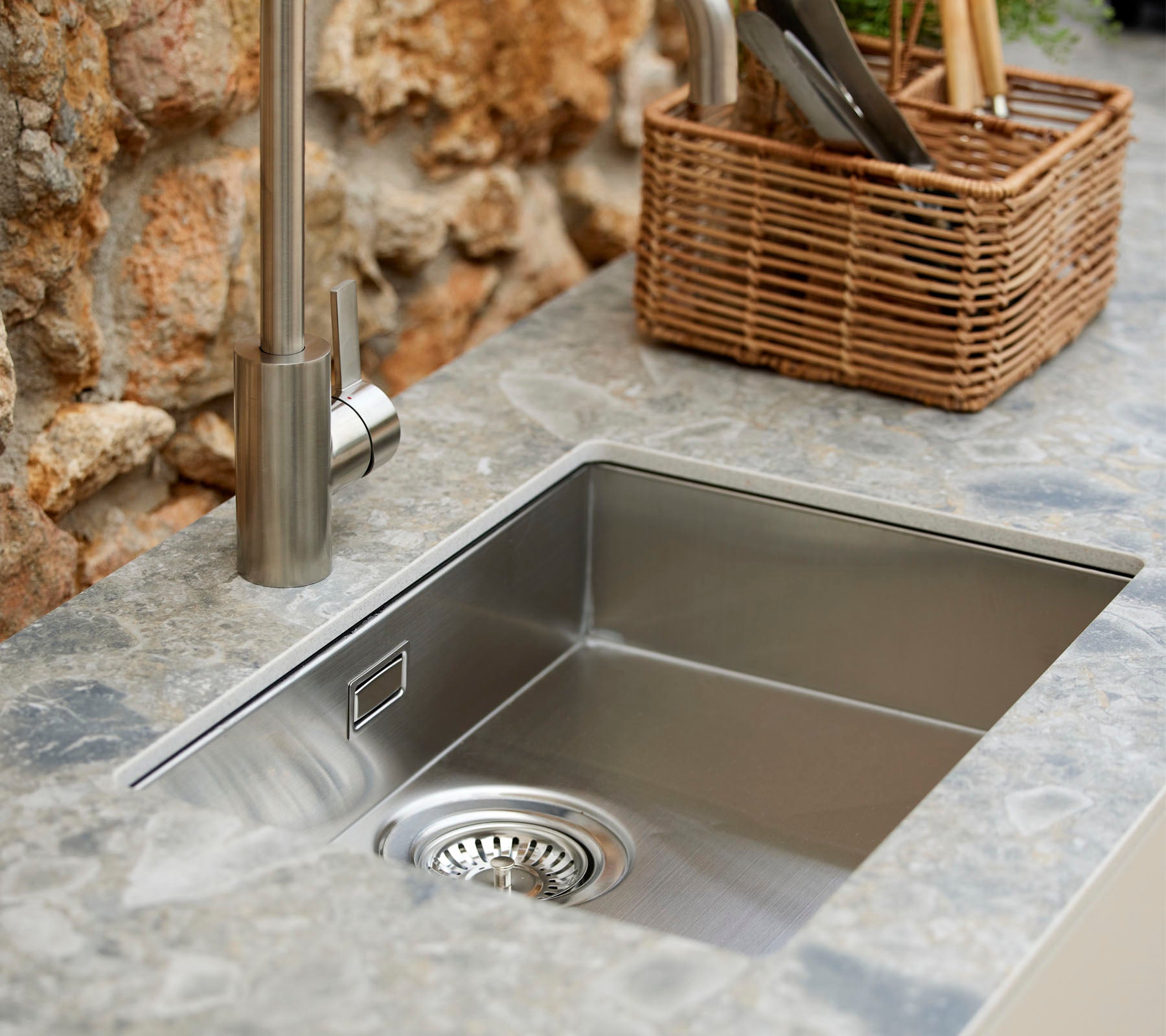 Stainless steel sink with a modern faucet, surrounded by a stone surface and a wicker basket holding kitchen utensils.