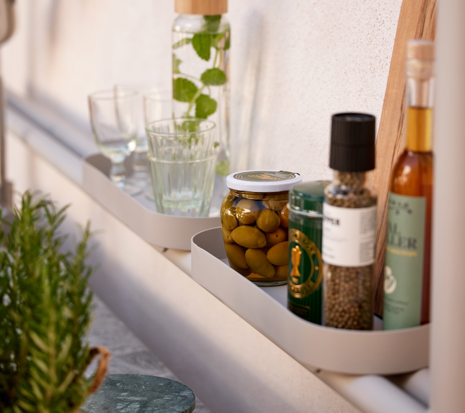 A shelf showcasing various kitchen items, including glasses, jars of olives, and bottles of oil and spices.