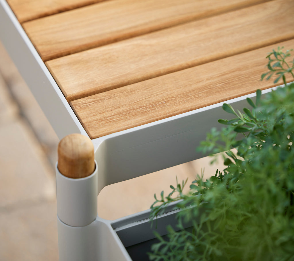 Teak slatted top bench with a white frame and green plants, featuring a rounded wooden stick detail.