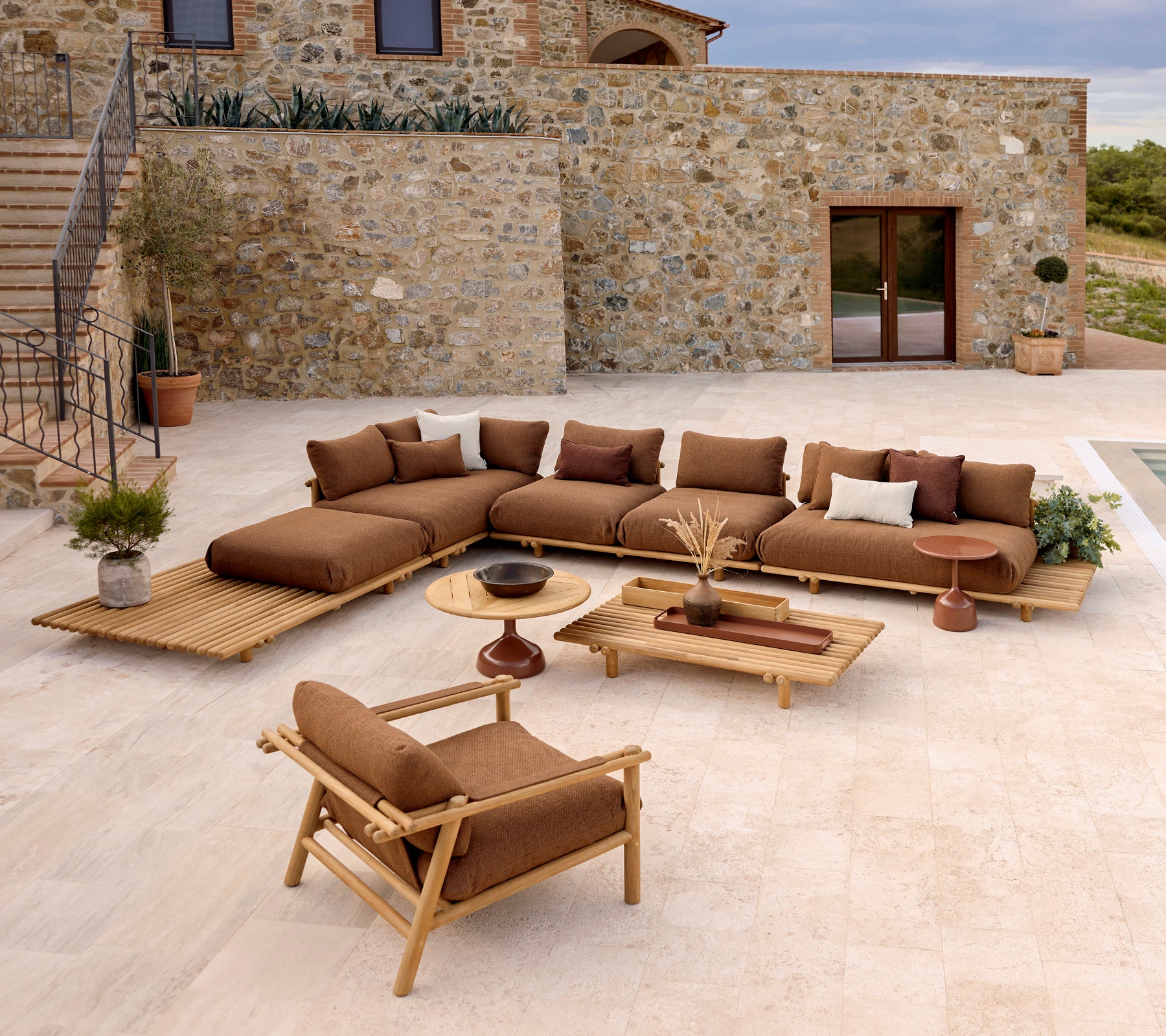 A stylish lounge area featuring a sectional sofa with plush cushions, a coffee table, and a teak lounge chair amidst a stone backdrop.