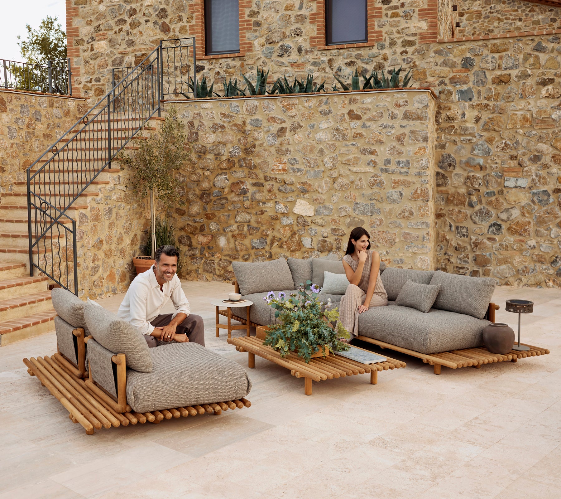 A stylish lounge area featuring teak furniture with plush gray cushions, accented by greenery and a modern coffee table.