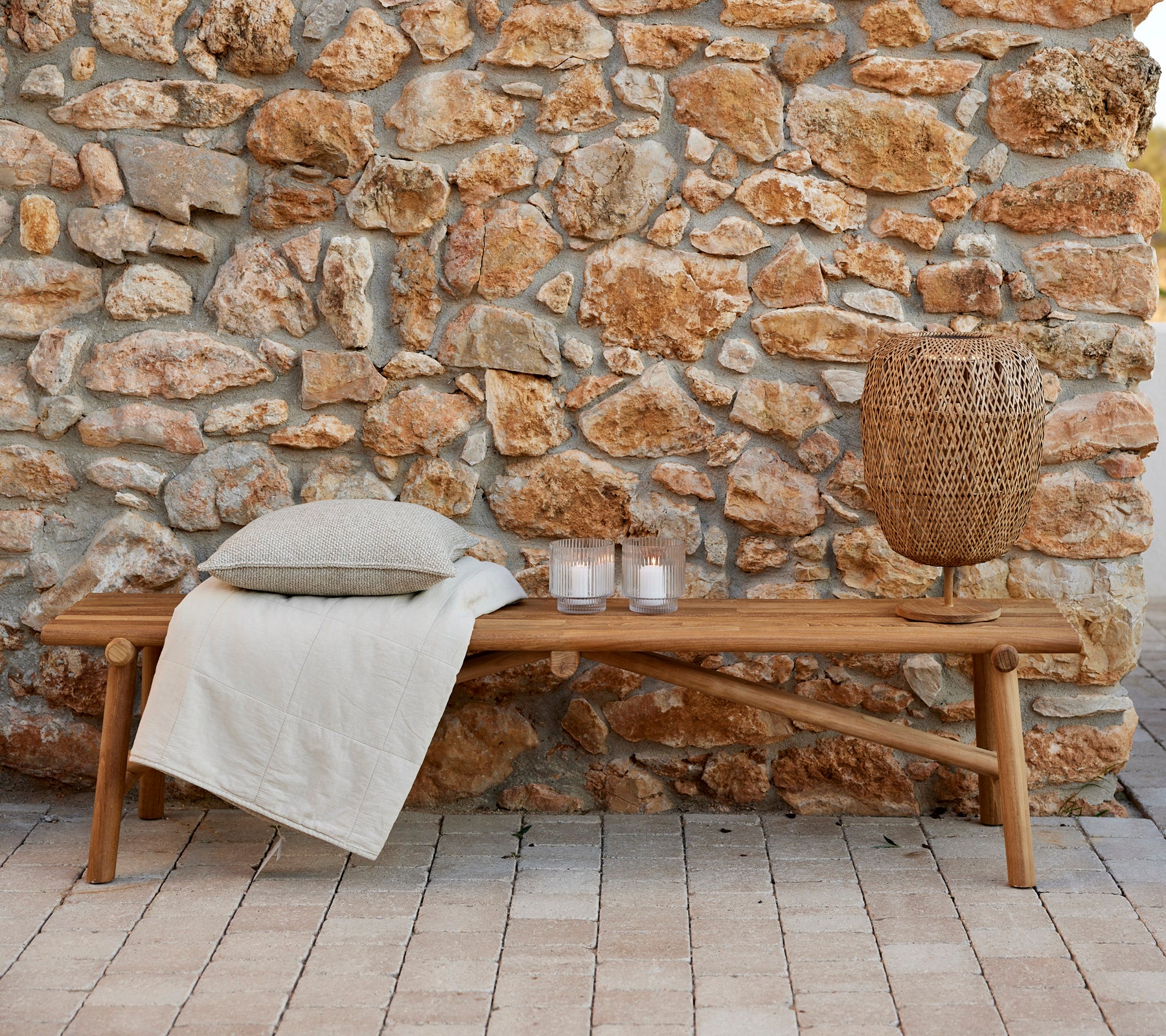 Teak bench with a cozy throw, decorative cushions, candles, and a woven lamp, set against a natural stone wall. Ideal for relaxation.