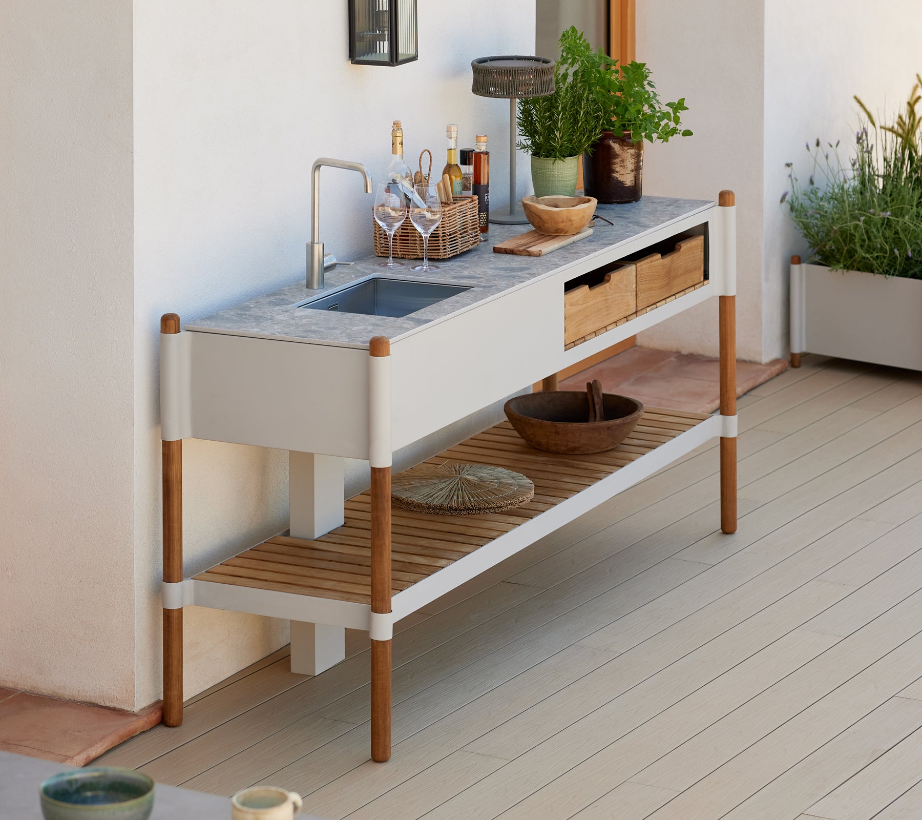 A modern, minimalist outdoor sink station with a granite countertop, wooden storage drawers, and decorative plant arrangements.